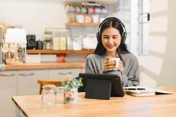 Young asian woman wearing headphones enjoys warm drink while using tablet in cozy kitchen setting. atmosphere is relaxed and inviting, perfect for leisure or work.