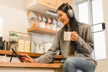 Wall Mural - Young woman relaxes in her modern kitchen enjoying music through headphones while sipping coffee and using a tablet, depicting a cozy lifestyle.