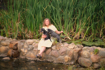 Wall Mural - Smiling woman sitting on a stone wall holding two small dogs near a pond with tall grass in the background. Outdoor lifestyle portrait.