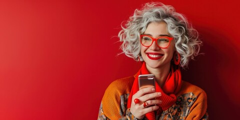 Cheerful woman with curly gray hair and red glasses holding a smartphone on a vibrant red background