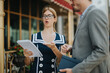 © qunica.com - Two young professionals are engaged in a lively discussion outside a modern office building, holding documents and expressing ideas passionately.