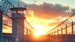 © Anoo - A prison entrance featuring a high fence with barbed wire and a guard tower under a captivating sunset sky