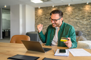 Satisfied businessman with credit card and laptop screaming and celebrating reward at desk in office