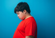 © idcreative.ddid - A side profile photograph of an overweight teen boy wearing a red t-shirt, standing with his head down, looking sad and dejected against a blue background