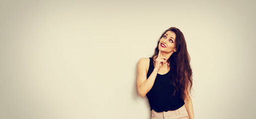 Beautiful positive fun makeup woman in black casual top looking up with finger under the face on empty isolated copy space studio background. Closeup