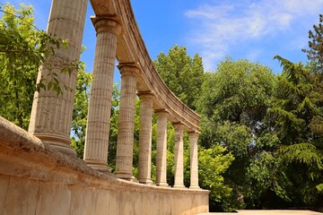 A semicircular rotunda, stone columns on a warm cream pedestal lined up against a backdrop of green trees
