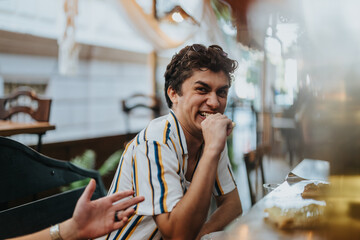 A cheerful young man in a striped shirt laughing at a cozy outdoor cafe setting. The casual and joyful atmosphere highlights friendship and enjoyment.