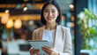 © IDNAF.std - Asian businesswoman is holding an iPad and smiling while standing in the office, wearing professional attire with her hands open to welcome you at work.