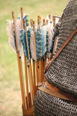 Closeup of blue and white fletching on the ends of the spines of arrows that are being held in a quiver on the back of a person wearing medieval chainmail armor.