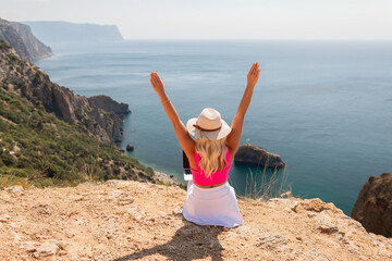 Wall Mural - A woman is sitting on a rocky hillside, wearing a pink top and a straw hat. She is looking out over the ocean, with her arms raised in the air. Concept of freedom and relaxation.