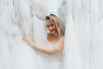 young beautiful caucasian blonde girl standing among wedding dresses, choosing wedding dress in bridal salon, preparing to wedding, choose wedding dress concept, copy space