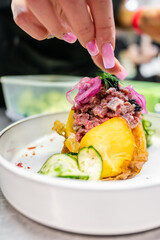 Poster - A close-up of a hand garnishing a dish with pink pickled onions. The plate features a stack of food with yellow and green elements, possibly vegetables, and a sprinkle of red spices