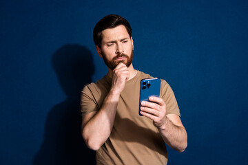 Aufkleber - Photo of young guy brunet in beige t shirt using smartphone touch chin reading news isolated on dark blue color background