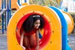 © Rakchanok - Young Afro hair elementary schoolgirl crawling through colorful tunnel on a playground, weekend carefree holiday in park. bright playground equipment and focus expression capture childhood exploration