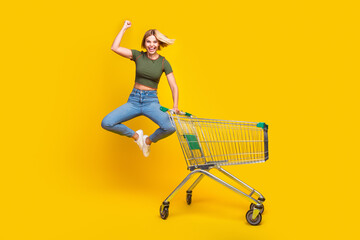 Wall Mural - Full size photo of ecstatic girl dressed khaki crop top hold shopping cart jump celebrate black friday isolated on yellow color background