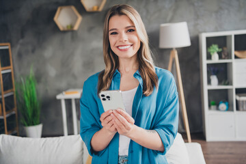 Photo of young confident business lady wearing blue stylish shirt using smartphone at her loft office selling renovated apartment for customers