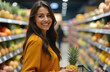 © SalenayaAlena - A young smiling woman holding a pineapple in her hands in a supermarket. Blurry shop shelves in the background.