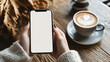 © Elnare - A woman's hands hold a smartphone with a blank screen, next to a cup of coffee with latte art.
