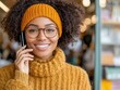 © MNFTs - Cheerful woman with curly hair wearing glasses and an orange sweater, talking on the phone in a cozy environment.