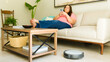 © AntonioDiaz - Plus size woman lounging on the sofa with her phone while a robotic vacuum cleans the living room