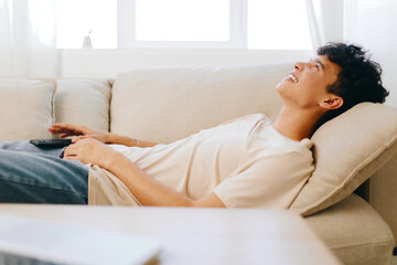 Relaxed young man lying on a sofa, enjoying leisure time, smiling with a remote control in hand, bright indoor setting with white curtains and natural light shining through