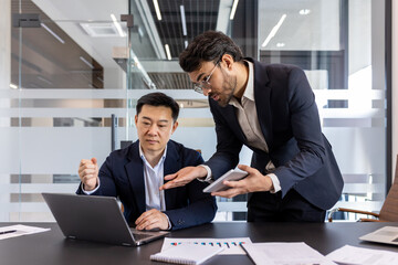 Two professionals engaged in strategic discussion in modern office. One seated using laptop and another showing information on phone. Concept of teamwork, collaboration, technology in business