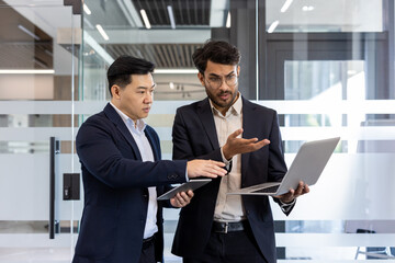 Wall Mural - Two businessmen engaged in discussion holding laptop and tablet in modern office. Displaying teamwork, communication in professional business environment. Emphasis on collaboration, technology