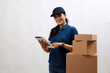 © ArvStd - Asian courier woman in blue standing with pile of cardboard box and ready to deliver to customer. Isolated on white background holding tablet pc computer