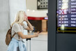 © Dejan - A woman with a backpack and suitcase examines the departure board in an railway station, checking her phone in hand while wearing casual travel attire, possibly anxious or curious.