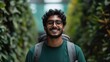 © familymedia - A cheerful man with glasses and a backpack smiles amid a lush green backdrop, exuding positivity and adventure, portraying a spirit of exploration and happiness.