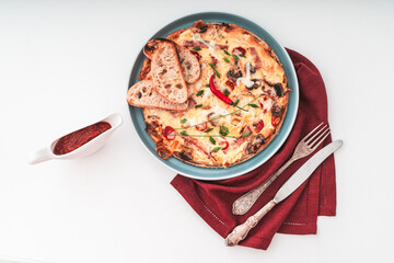 Frittata or Italian omelette with ham, tomatoes, cheese on a blue plate with slices of toasted bread and red sauce with a burgundy napkin and cutlery on a white table. Selected focus. Copy space