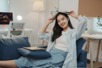 Young woman is taking a break from working at home, relaxing on the sofa with a book on her head and smiling