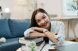 © amnaj - Young woman is smiling while leaning on a stack of books at a table in her home