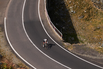 Aerial view of a cyclist riding on a winding mountain road. Road is lined with white lines and the cyclist is wearing a white jersey. Background is a lush green forest.Cycling in Carpathian Mountains