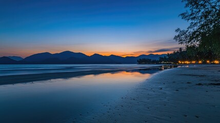 A tranquil beach scene at dusk with a clear reflection of the mountain range in the still water.