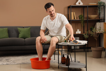 Wall Mural - Handsome young happy man with tools keeping his legs in foot basin and doing pedicure at home