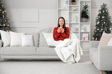 Sticker - Happy young woman with cup of cacao sitting on sofa near Christmas tree at home