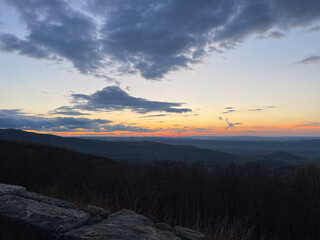 An orange and yellow sunset lights up the sky from Skyline Drive in Shenandoah National Park, casting a warm glow over the Blue Ridge Mountains. The vibrant colors create a breathtaking end to the day