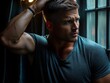 © TesaI - A handsome man with short hair poses thoughtfully by a window, running his hand through his hair, illuminated by soft, warm light