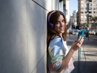 A young woman with headphones smiles while listening to music on her smartphone, leaning against a wall in a vibrant urban setting during the afternoon.