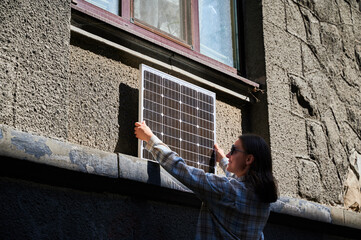 Woman holding photovoltaic solar panel in front of historical building, wearing plaid shirt and sunglasses. Concept of integration of sustainable renewable energy sources into old architecture.