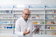 © PhugunStock - Asian professional male pharmacist using tablet checking drug list holding pill bottle Pharmacist checking stock in pharmacy on shelf looking at camera charming smile
