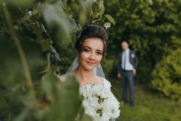 Wall Mural - A bride and groom are posing for a picture in a lush green field. The bride is holding a bouquet of white flowers, and the groom is wearing a suit. Scene is romantic and happy