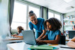 © Maskot - Female teacher assisting curly hair girl studying on laptop while sitting near desk in classroom at elementary school