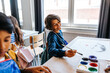 © Maskot - Happy boy painting with paintbrush while sitting near desk in art class at elementary school