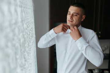 A man in a white shirt is getting dressed in front of a window. He is wearing a tie and he is in a good mood