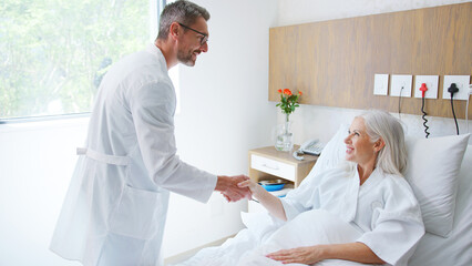 Wall Mural - Mature Male Doctor Or Surgeon Shaking Hands With Mature Female Patient Lying In Hospital Bed