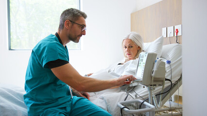 Male Nurse Taking Mature Female Patients Blood Pressure In Hospital Bed With Automated Machine