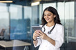 © Liubomir - Smiling female doctor stands in modern office using digital tablet. Stethoscope around neck suggests healthcare setting. Image conveys professionalism, technology integration, and positive work