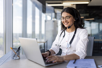 Wall Mural - Confident female doctor wearing glasses and headset engaged in online consultation using laptop. Stethoscope around neck signifies professional medical setting.